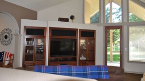 A living room with a large flat-screen TV, wooden entertainment center, and a view through a glass door. - Perfect Moments B&B and Event Venue (Nampa)