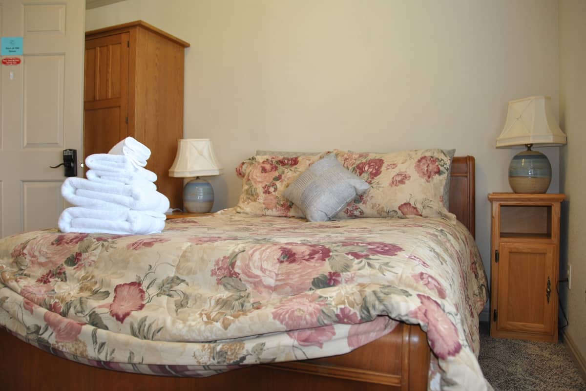 A neatly made bed with floral bedding, a stack of folded towels, a wooden wardrobe, a bedside lamp, and a small table with a lamp.