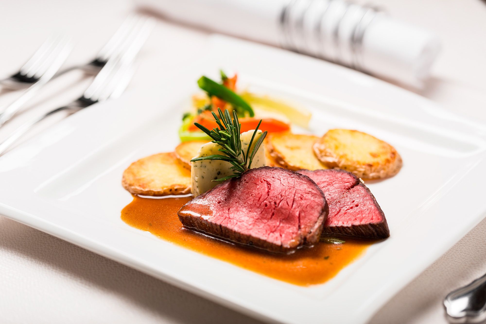 A white plate with slices of medium-rare steak, a small herb garnish, and a side of potato wedges.