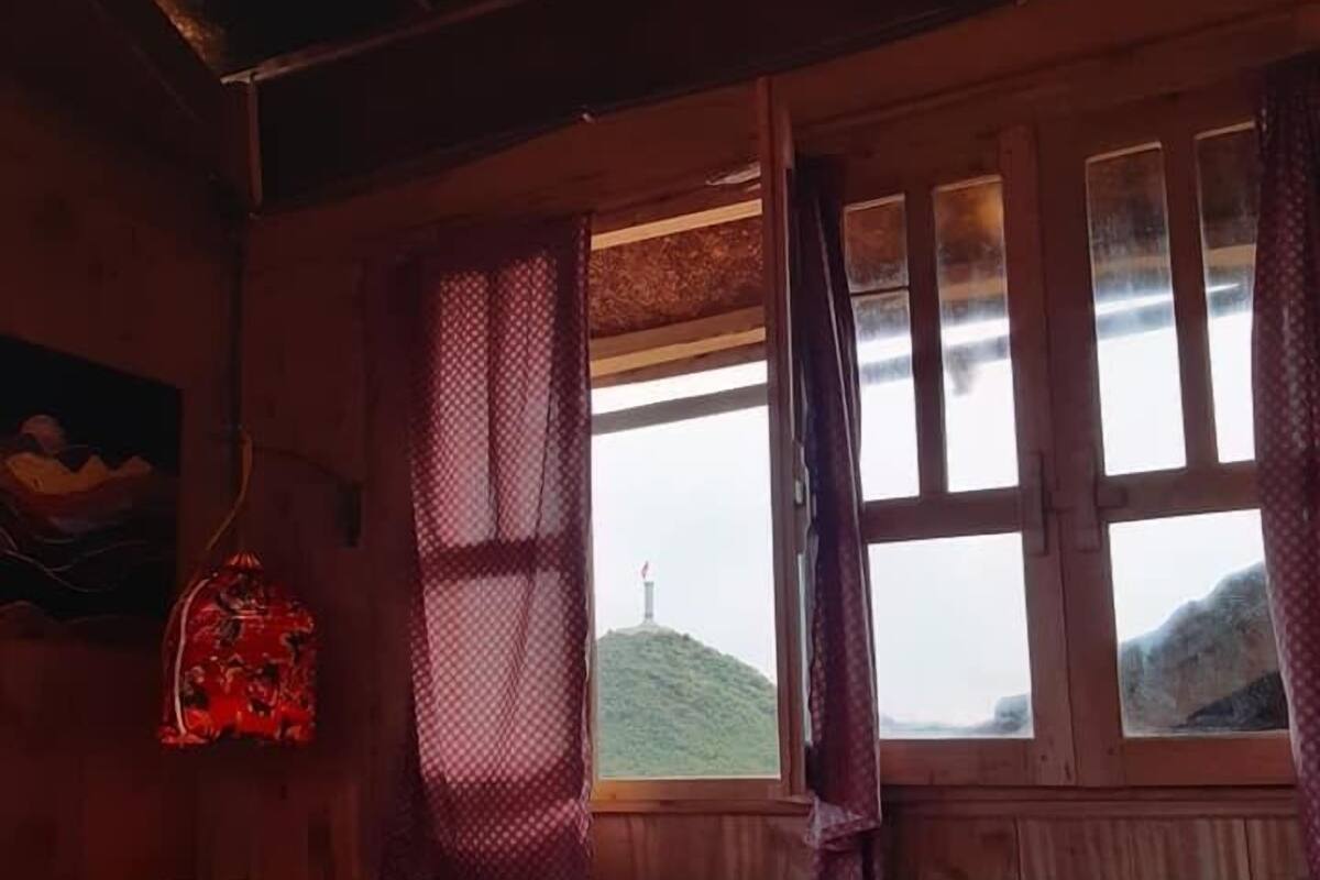 A room with a bed, a window with curtains, a wall-mounted lamp, and a view of a hill.