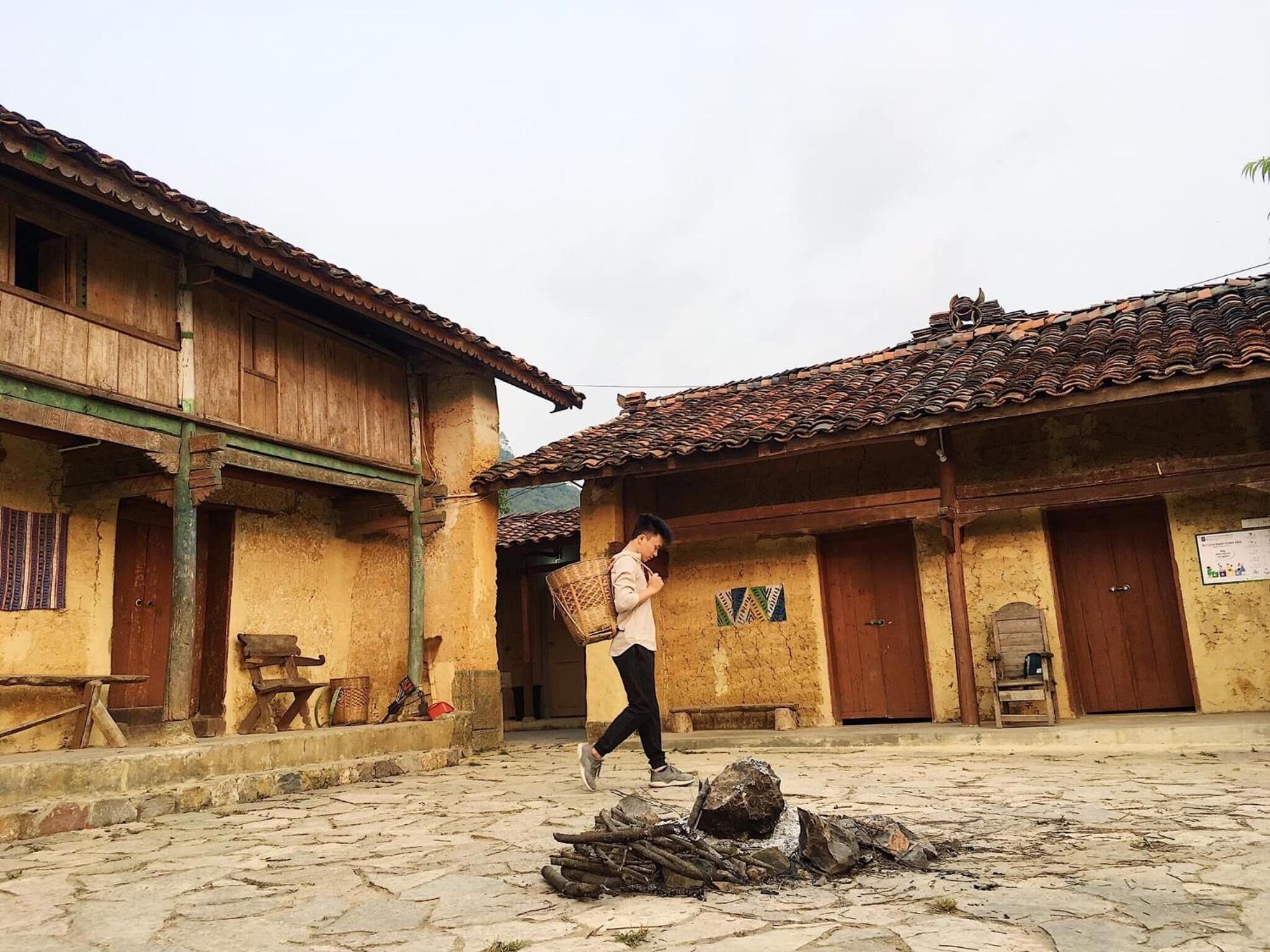 A rustic courtyard with traditional wooden buildings, a stone-paved ground, and a person carrying a basket.