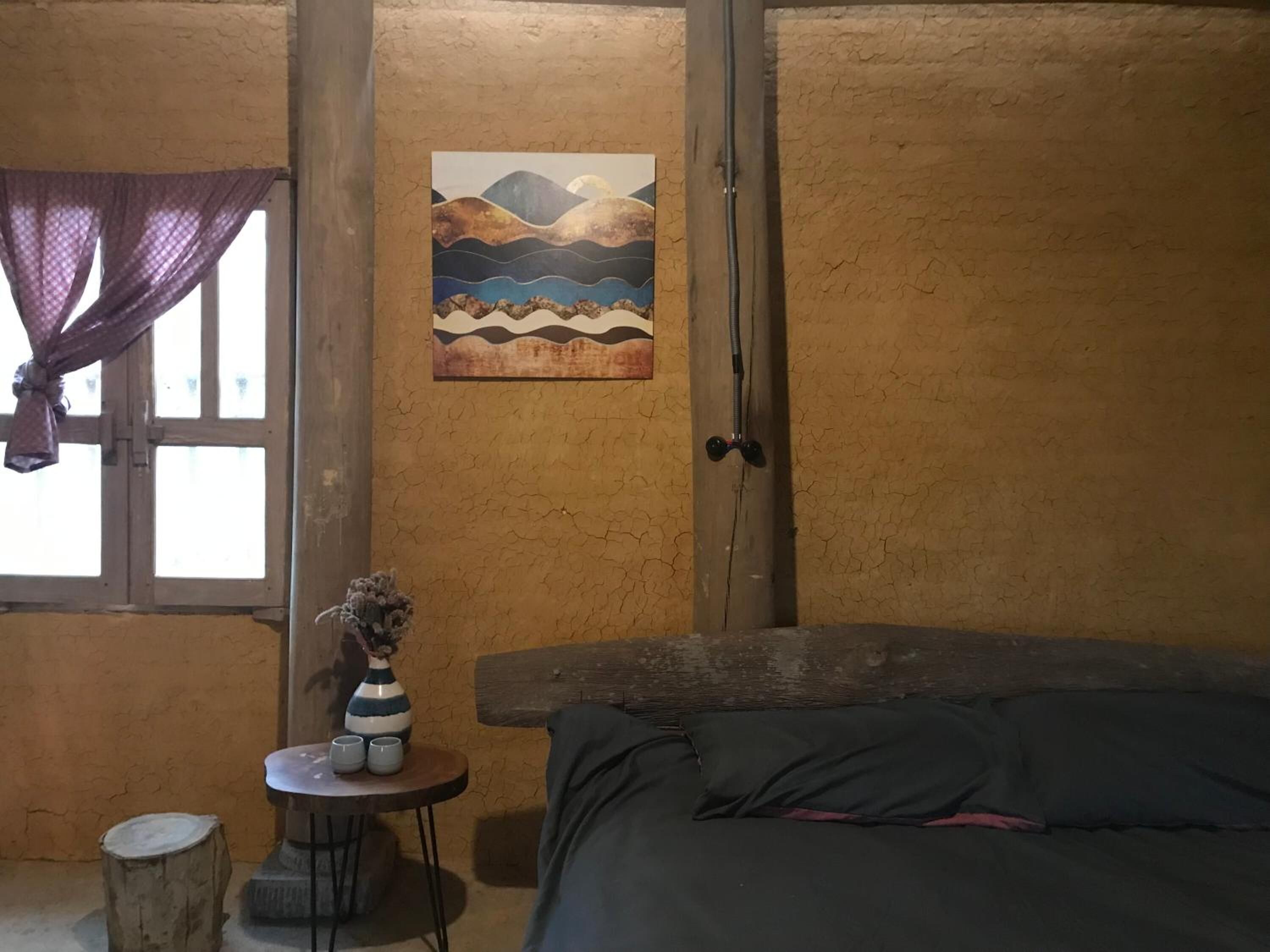 A room with a bed, a small table with a vase and cups, a window with a curtain, and a painting on the wall.