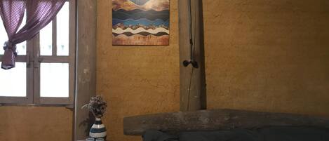 A room with a bed, a small table with a vase and cups, a window with a curtain, and a painting on the wall.