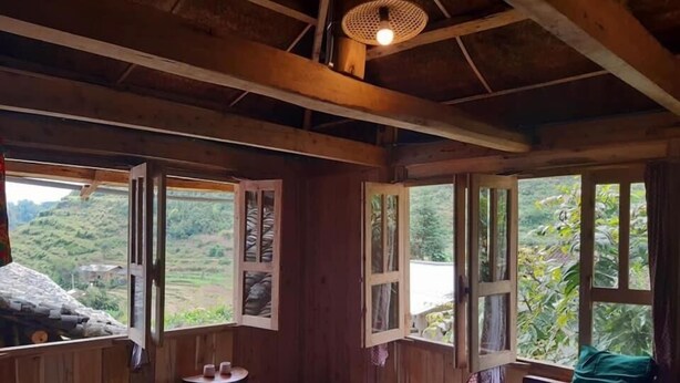 Una baita di legno con un letto, un tavolino e finestre aperte su un paesaggio verde.