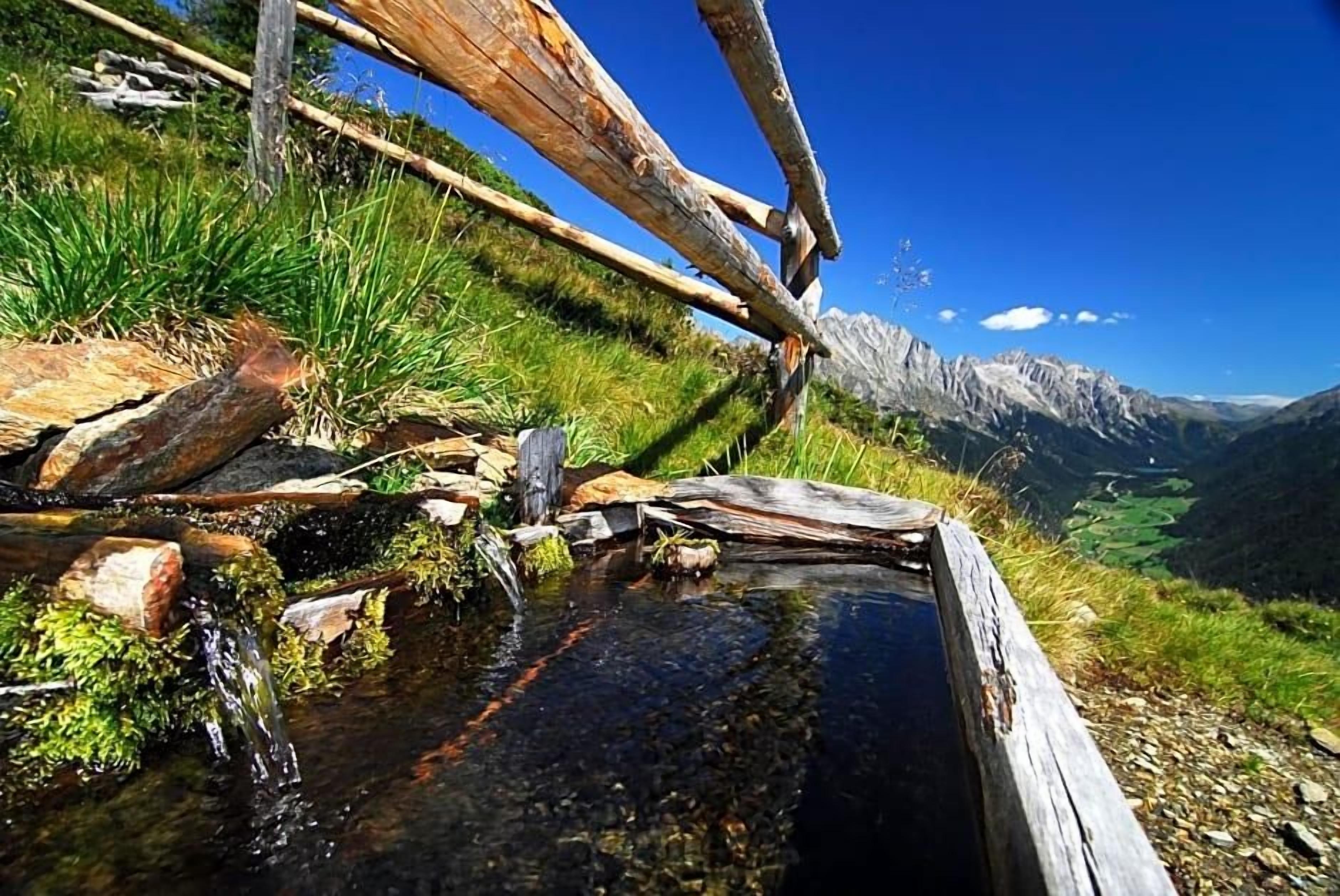 Une construction rustique en bois, avec un ruisseau qui serpente entre les rochers, le tout entouré de verdure et dominé par un paysage montagneux.
