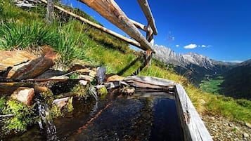 Une construction rustique en bois, avec un ruisseau qui serpente entre les rochers, le tout entouré de verdure et dominé par un paysage montagneux.
