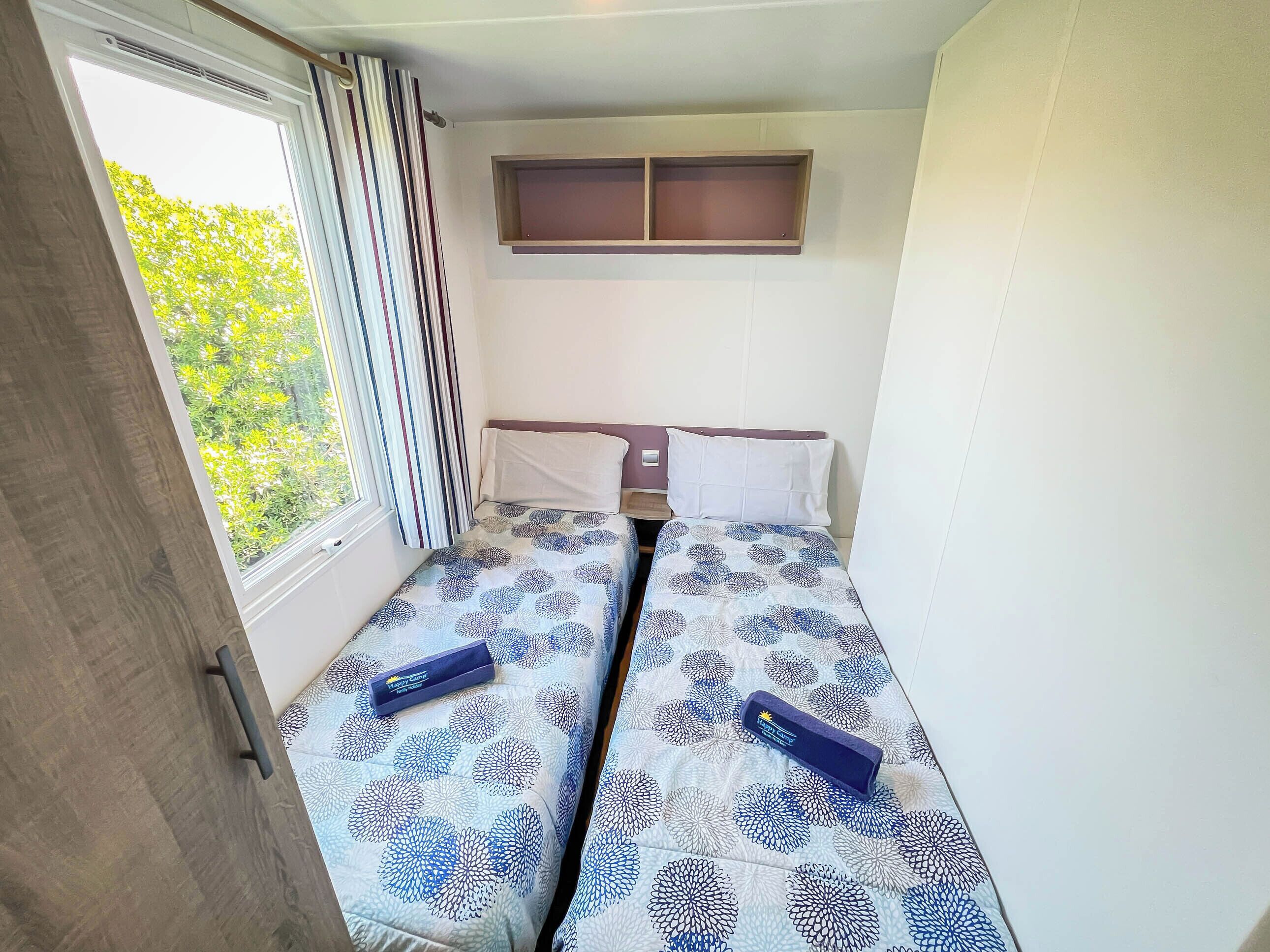 Una habitación pequeña con cama individual, ventana, cortina, mesita de noche y estante sobre la cama.