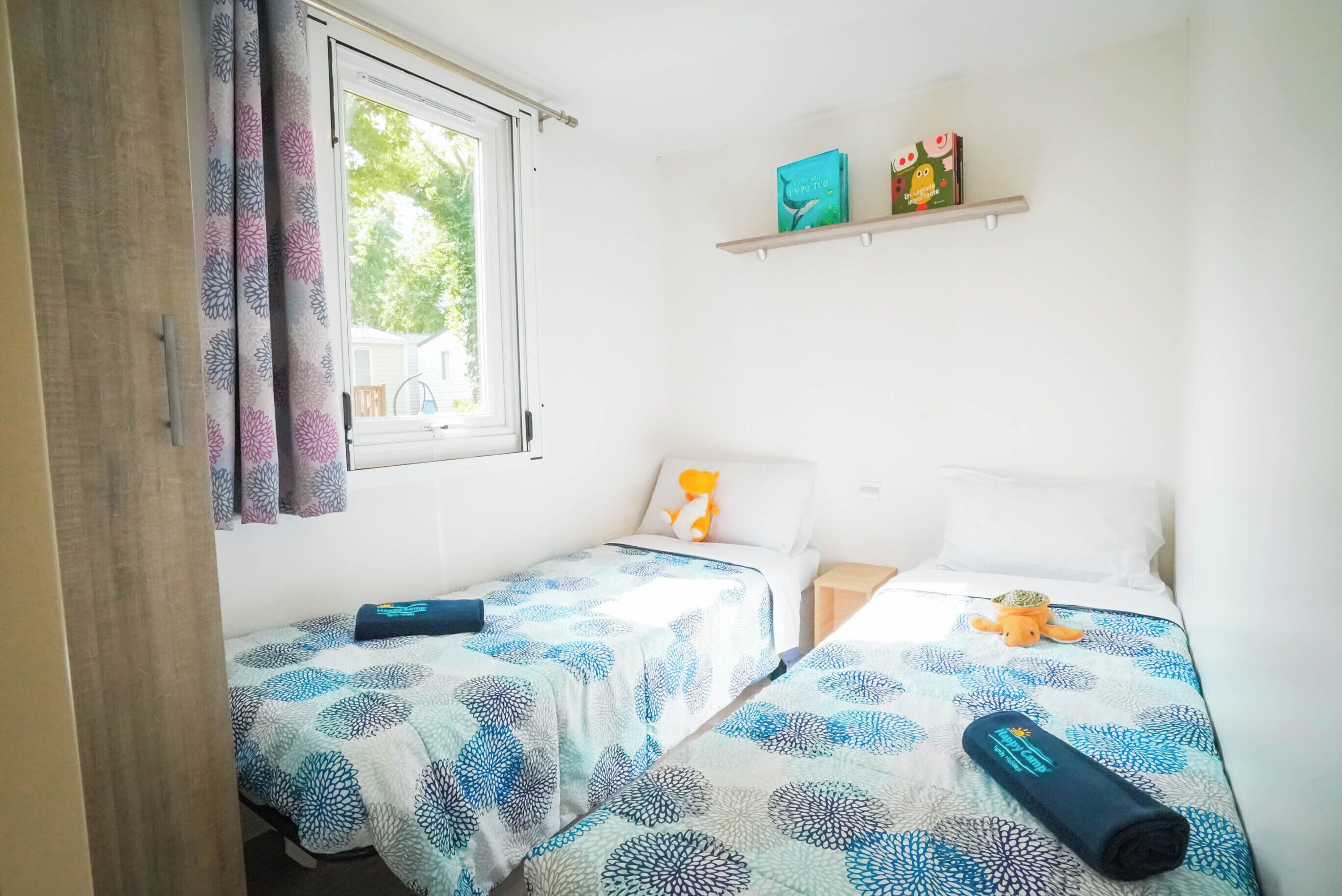 Um quarto pequeno e limpo, com duas camas de solteiro, uma janela com cortinas, uma estante com livros e uma planta, e uma mesinha.