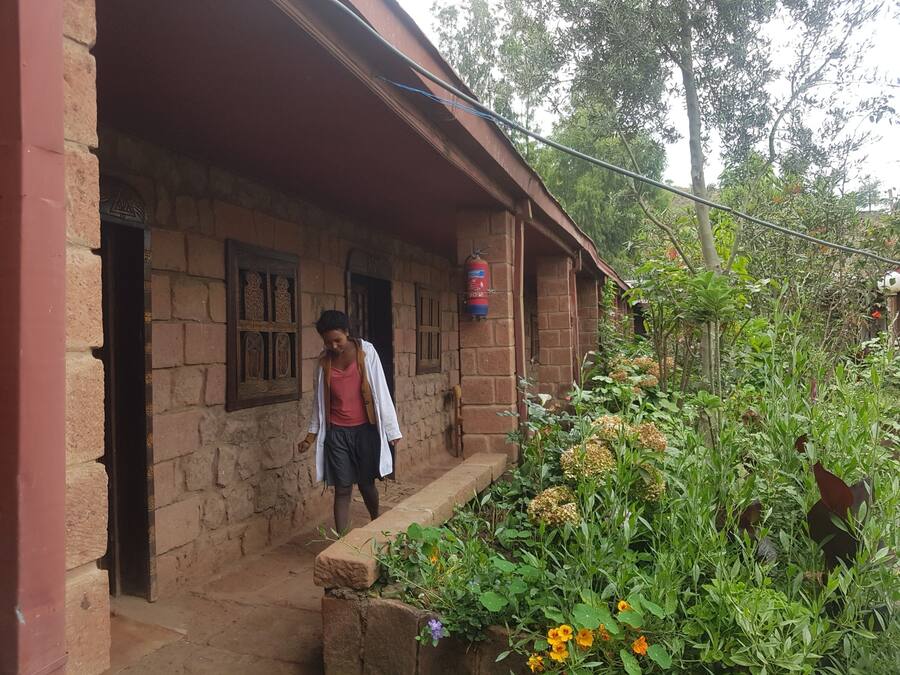A rustic brick building with a porch, a fire extinguisher mounted on the wall, and a garden with various plants and flowers.