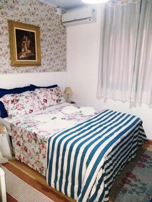 Un dormitorio con una colcha de estampado floral, una manta a rayas, un cuadro en la pared y un equipo de aire acondicionado.
