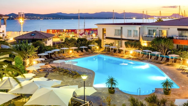 A swimming pool with lounge chairs and umbrellas, surrounded by buildings with balconies, and a view of the sea and mountains. - Best Western La Marina (Saint-Raphael)