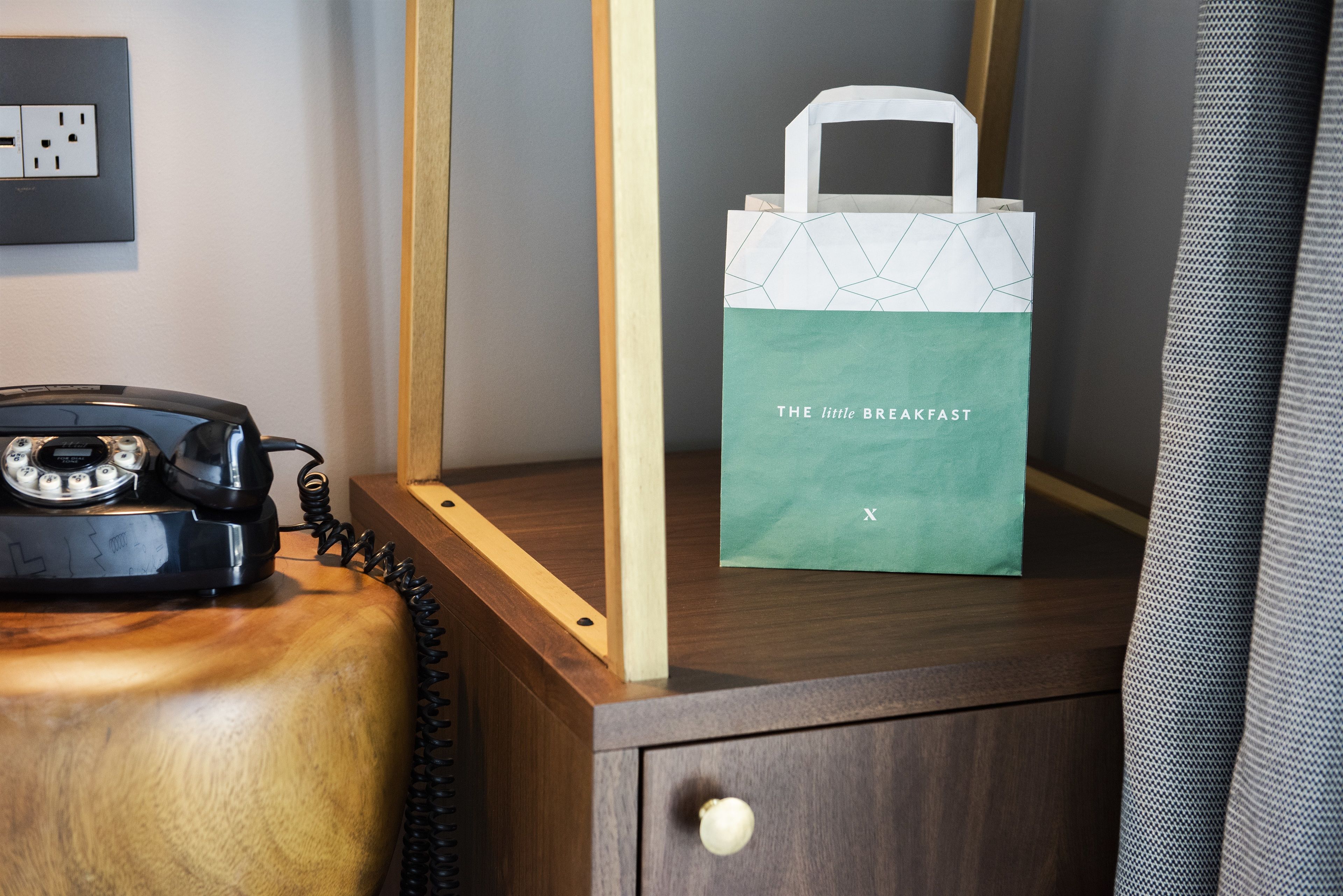 Una mesita de noche de madera con un teléfono giratorio antiguo, un bolso de mano azul aguamarina y blanco con la etiqueta "The Little Breakfast", y un tocador de madera.