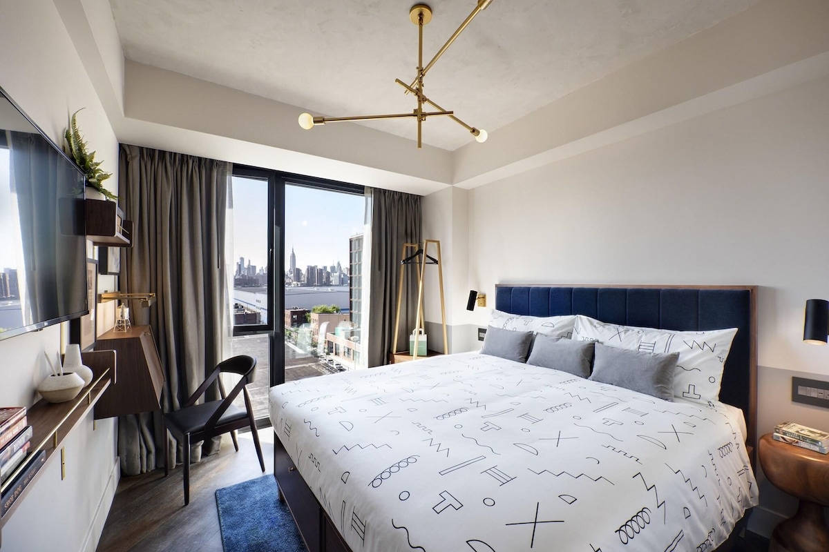 A modern hotel room with a large bed, a desk with a chair, a TV, and a view of the city.