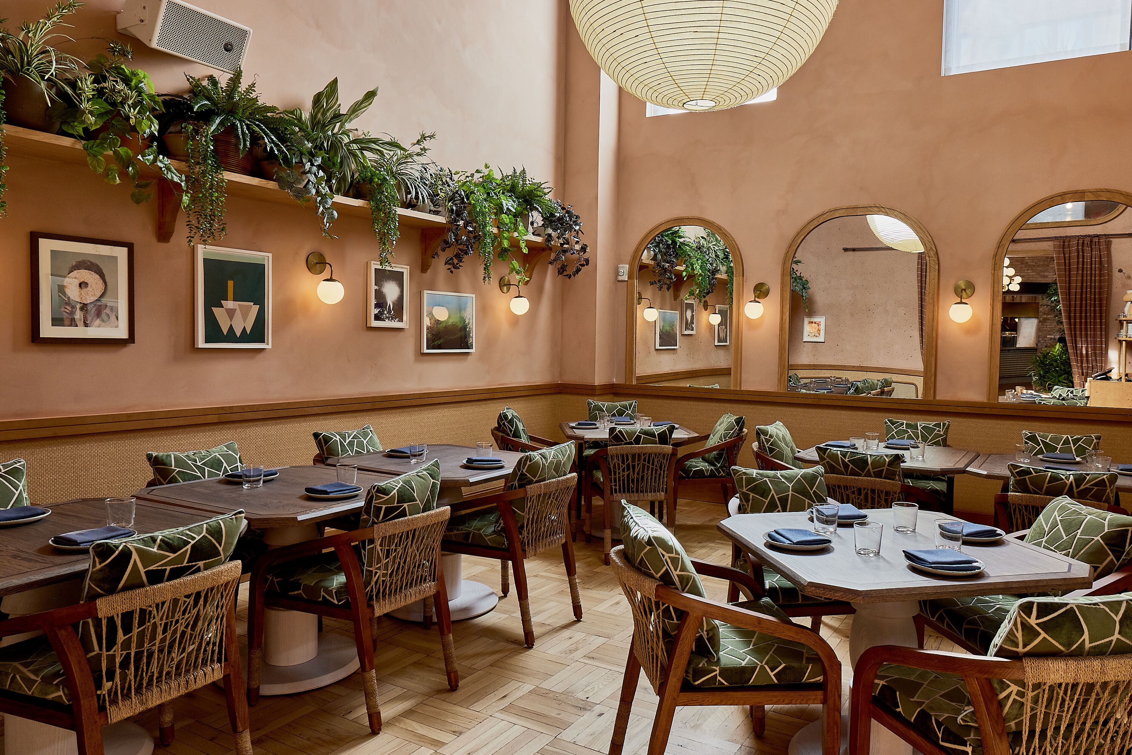 Een restaurant met houten tafels en stoelen, groene kussens met een patroon, wandplanten, ingelijste foto's en hanglampen.