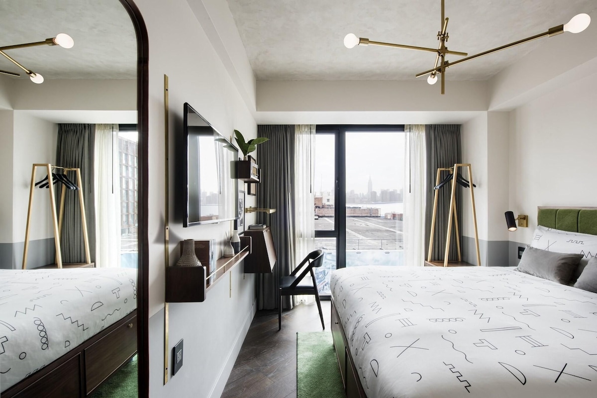 A modern hotel room with two beds, a desk, and a large window offering a city view.