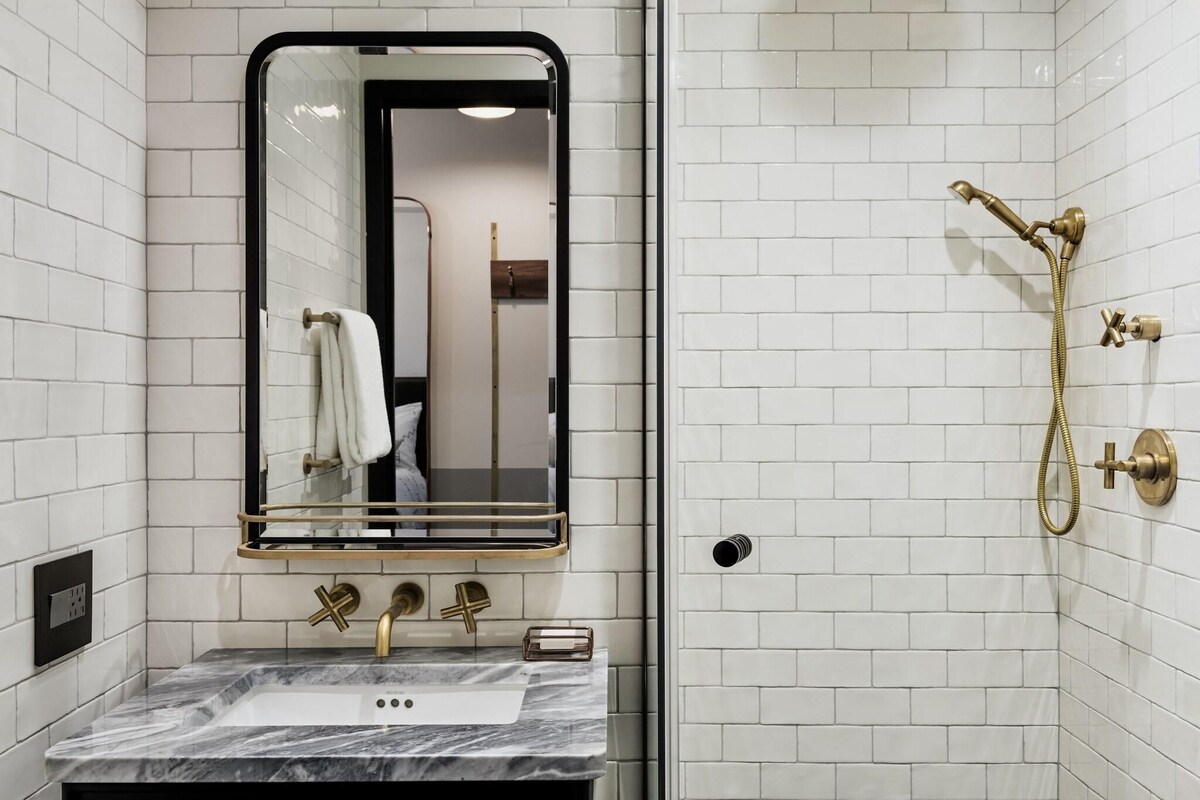 A bathroom with white tiled walls, a marble sink, a gold showerhead, and a mirror with a gold frame.