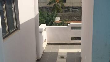 Um corredor estreito com azulejos, paredes brancas e uma janela à esquerda. Uma palmeira e um edifício são visíveis ao fundo.