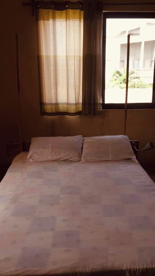 A bed with a checkered bedspread, a window with a curtain, and a visible electrical outlet on the wall.