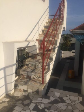 Una escalera con barandilla roja que lleva a un nivel superior. Los escalones están hechos de losas de piedra, y hay un suelo de baldosas en la parte inferior.