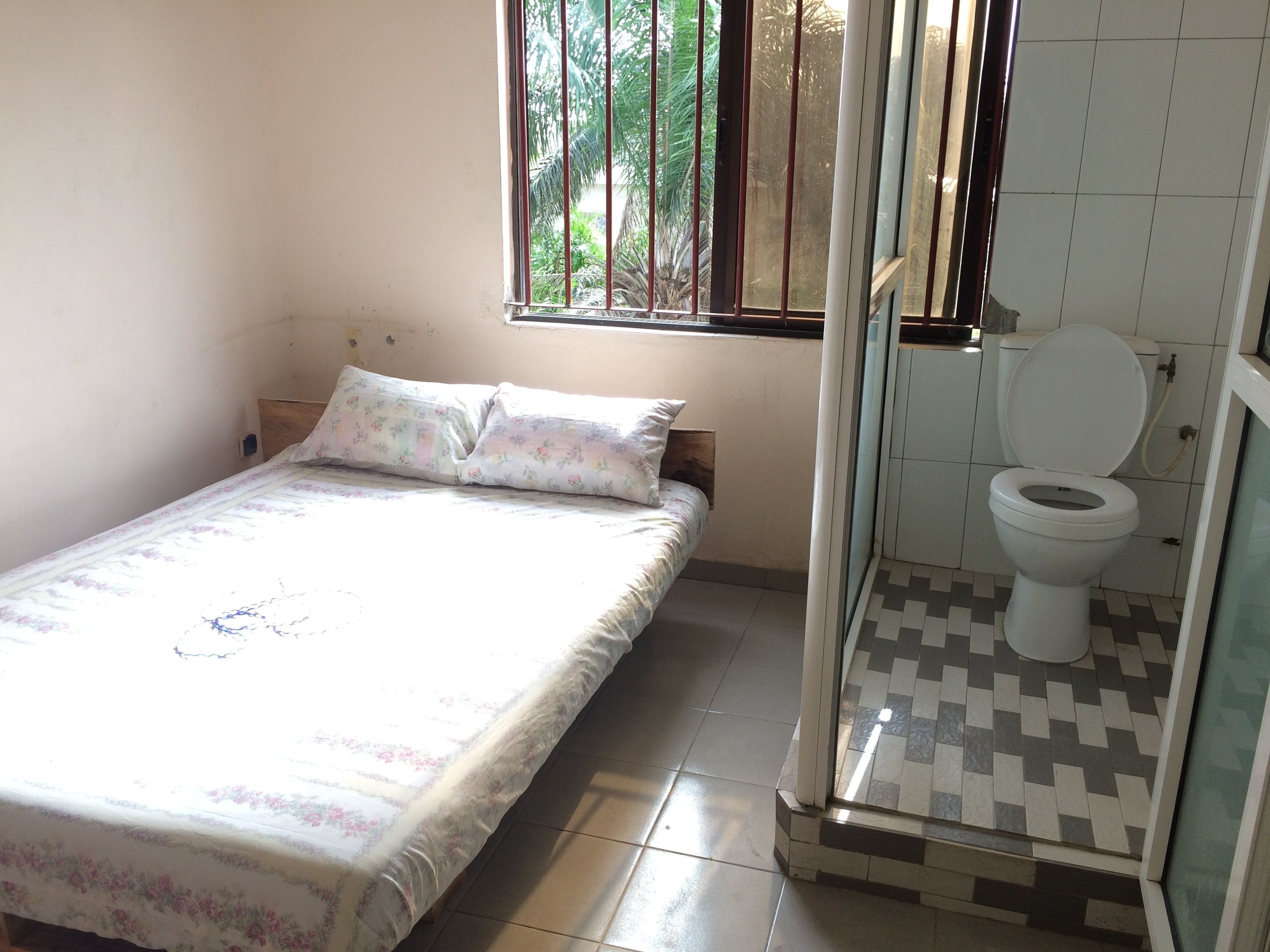 A small, single-bed room with a window, a toilet, and a tiled floor.