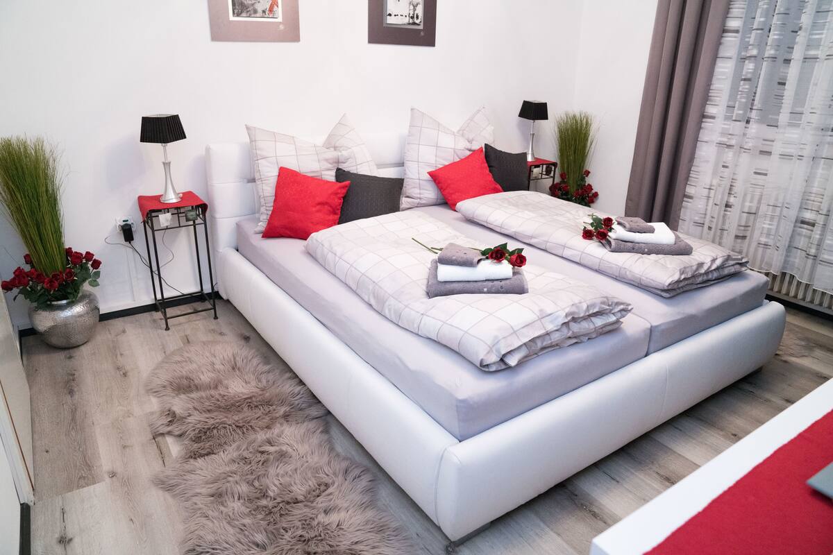 A modern bedroom with a large bed, two bedside tables with lamps, a fluffy rug, and a vase with red flowers.