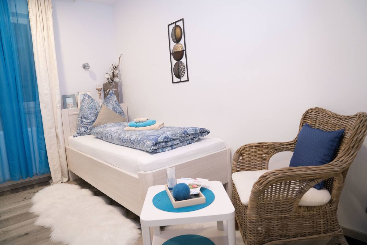 A bedroom with a bed, a wicker chair, a small table, and a wall art piece.