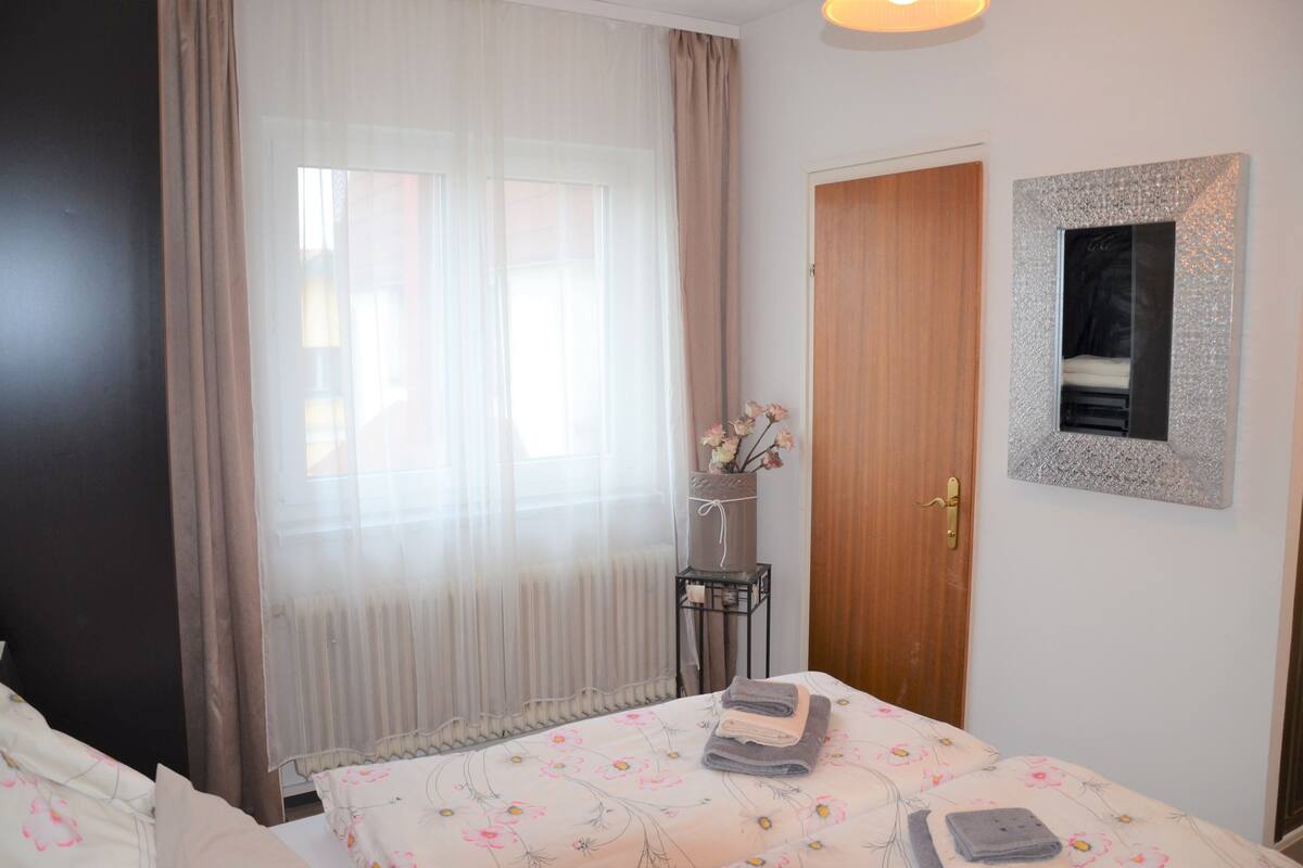 A hotel room with a bed, a wooden door, a mirror, and a window with sheer curtains.