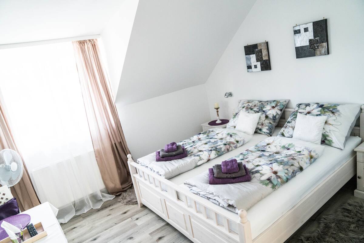 A bedroom with a white bed, floral bedding, and a small bedside table with a lamp.