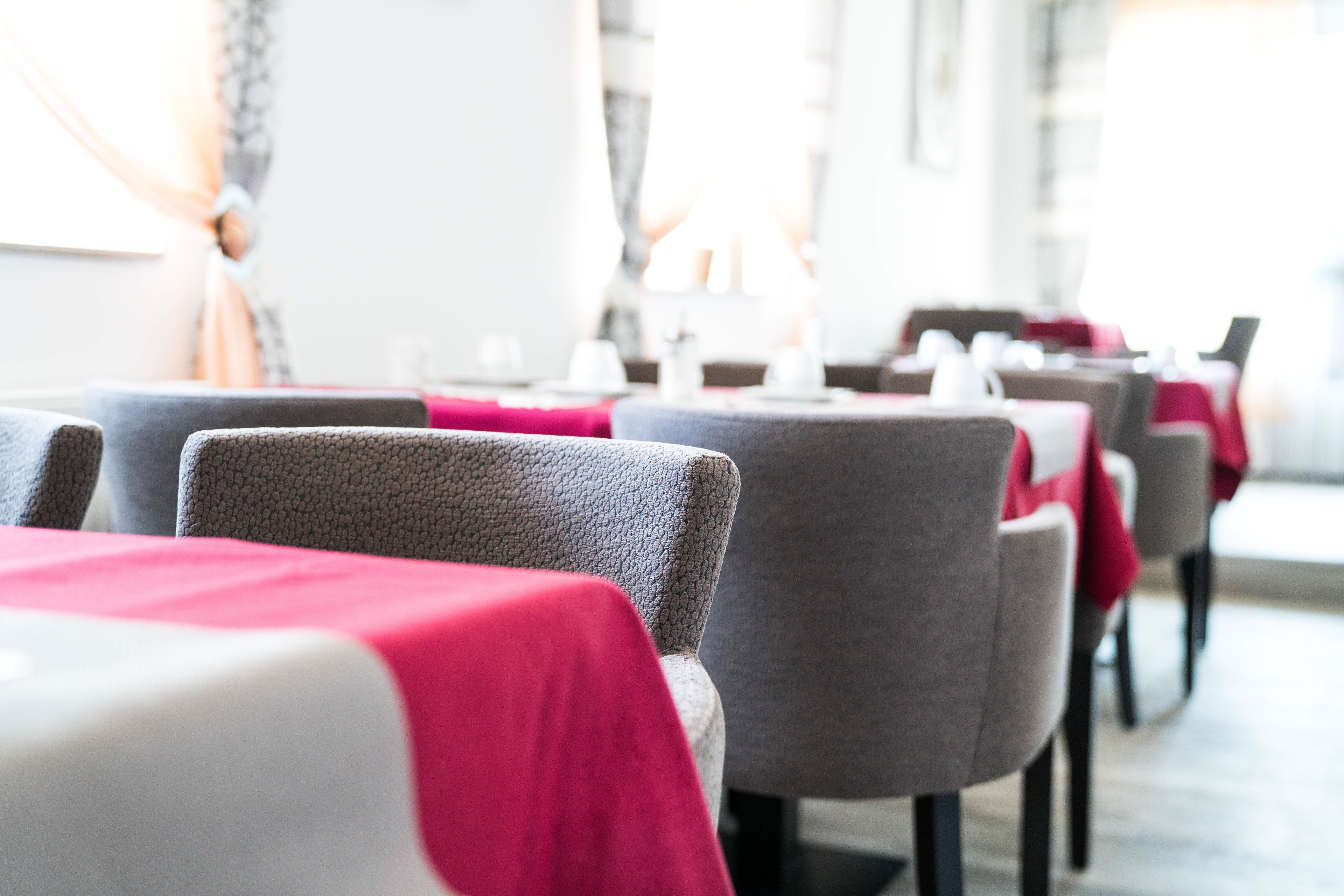 A row of dining tables with red and white tablecloths, grey upholstered chairs, and a whiteboard in the background.