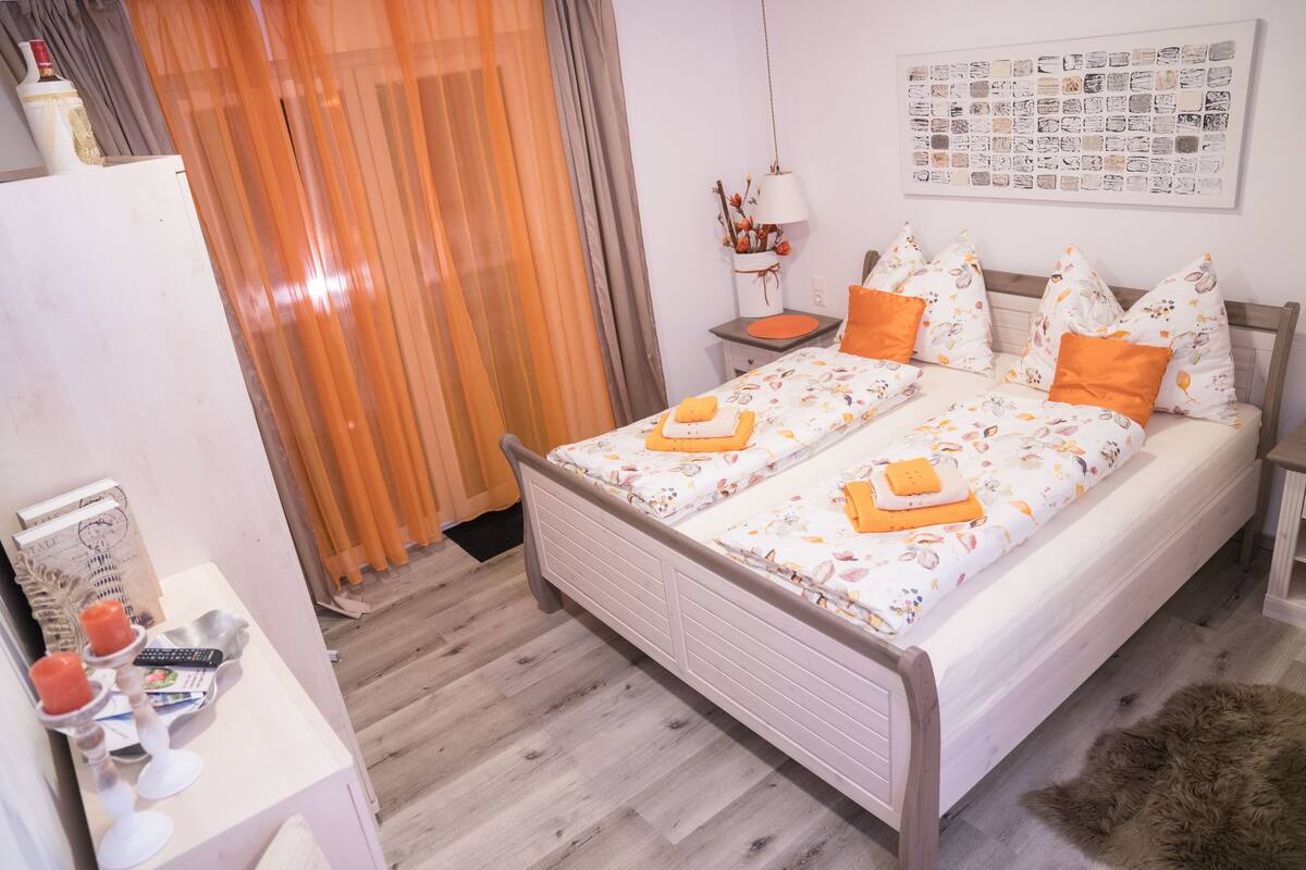 A bedroom with a double bed, a bedside table, a nightstand, a lamp, a framed picture on the wall, and a window with curtains.