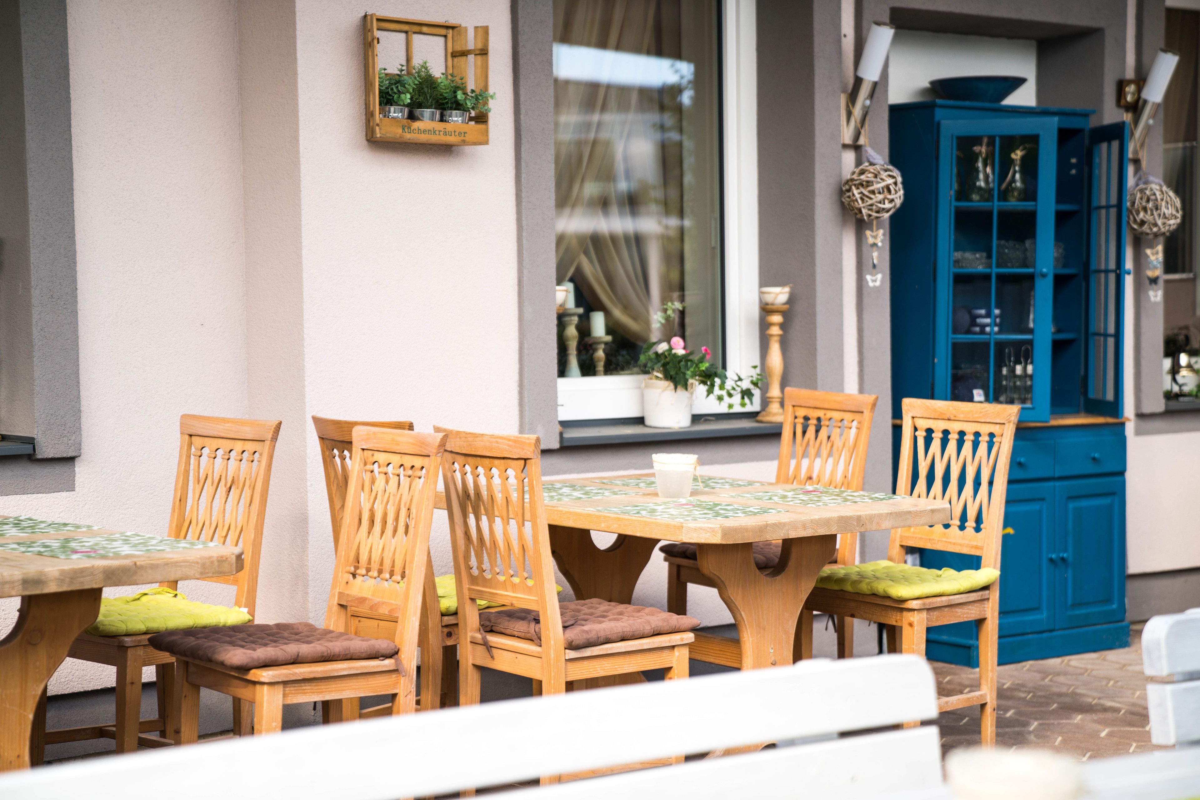 Un área de comedor al aire libre con mesas y sillas de madera, un armario azul y una ventana con cortinas.