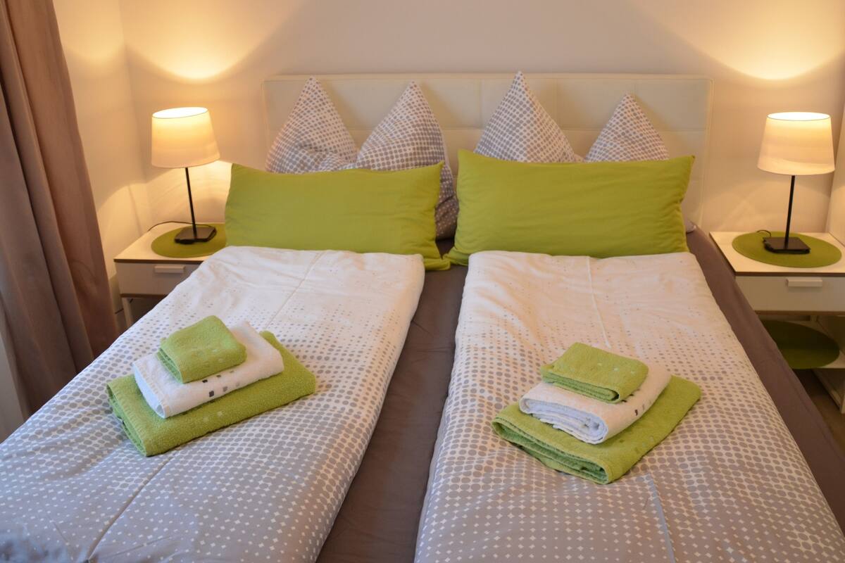 A hotel room with two beds, each with a green pillow and a folded towel. There are two bedside lamps on each nightstand.