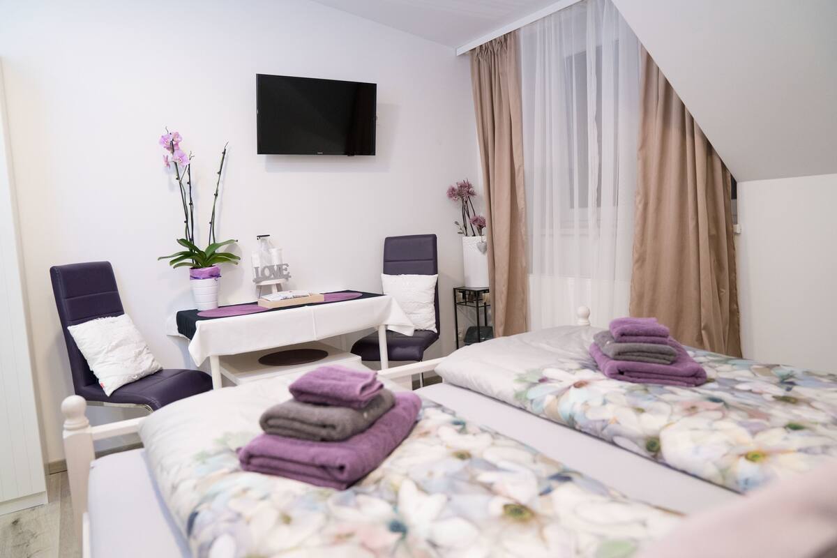 A bedroom with two beds, a desk, chairs, a TV, and a window with curtains.