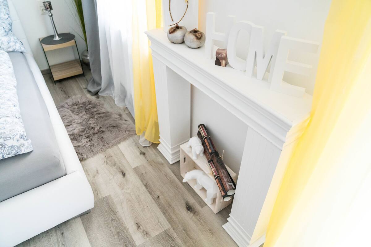 A bedroom with a bed, a white fireplace, and a small bedside table with a lamp.