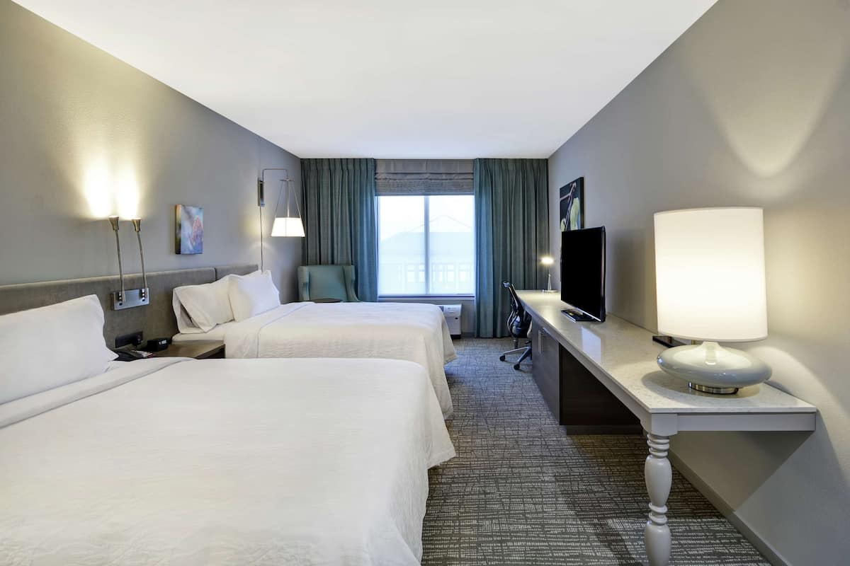 A hotel room with two beds, a desk with a TV, a lamp, and a view of the city.
