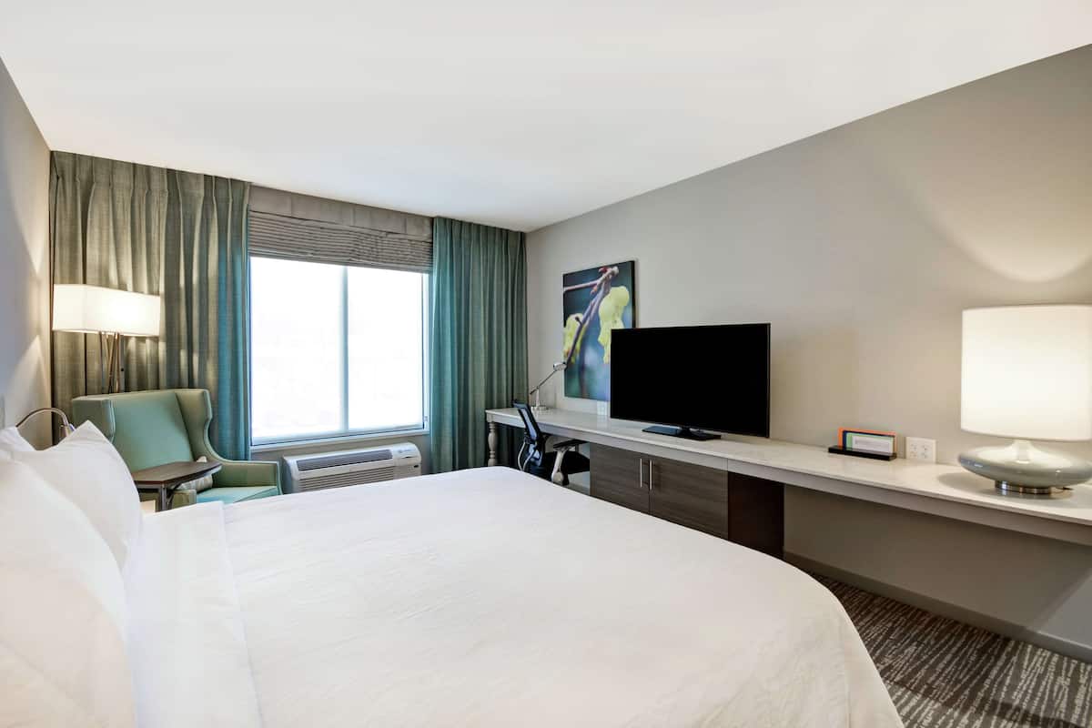 A hotel room with a large bed, a desk with a flat-screen TV, a chair, and a window with curtains.