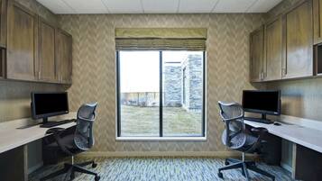 Una habitación con dos escritorios, cada uno con monitor, teclado y silla. Hay un ventanal con vistas a un edificio y una zona ajardinada. La habitación tiene armarios de madera y alfombra estampada.