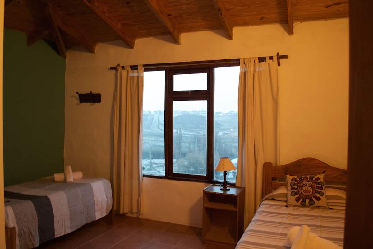 A room with two beds, a window with curtains, a nightstand with a lamp, and a view of the outside.