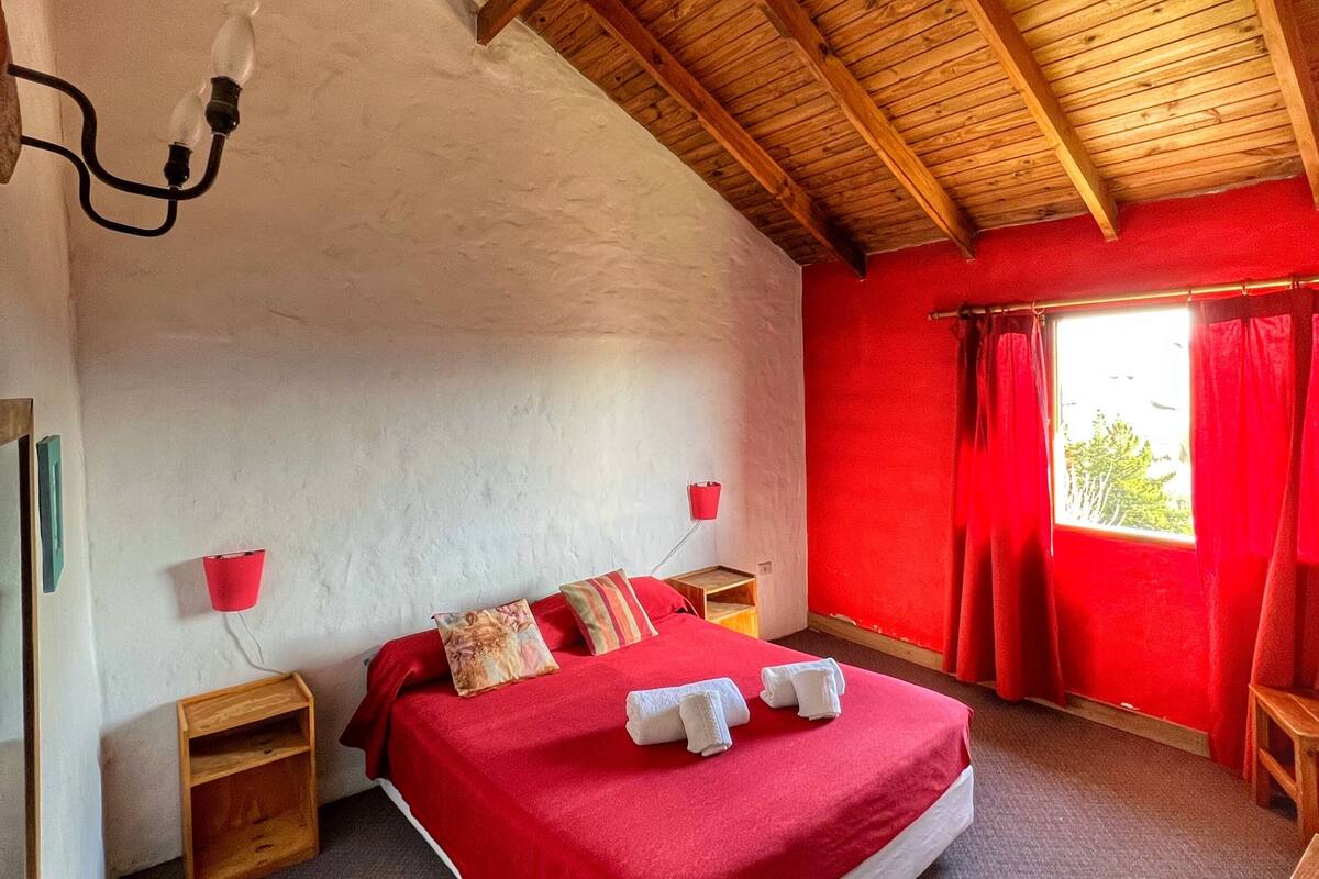 A bedroom with a wooden ceiling, a red bedspread, two pillows, a nightstand, a red lamp, and a window with red curtains.