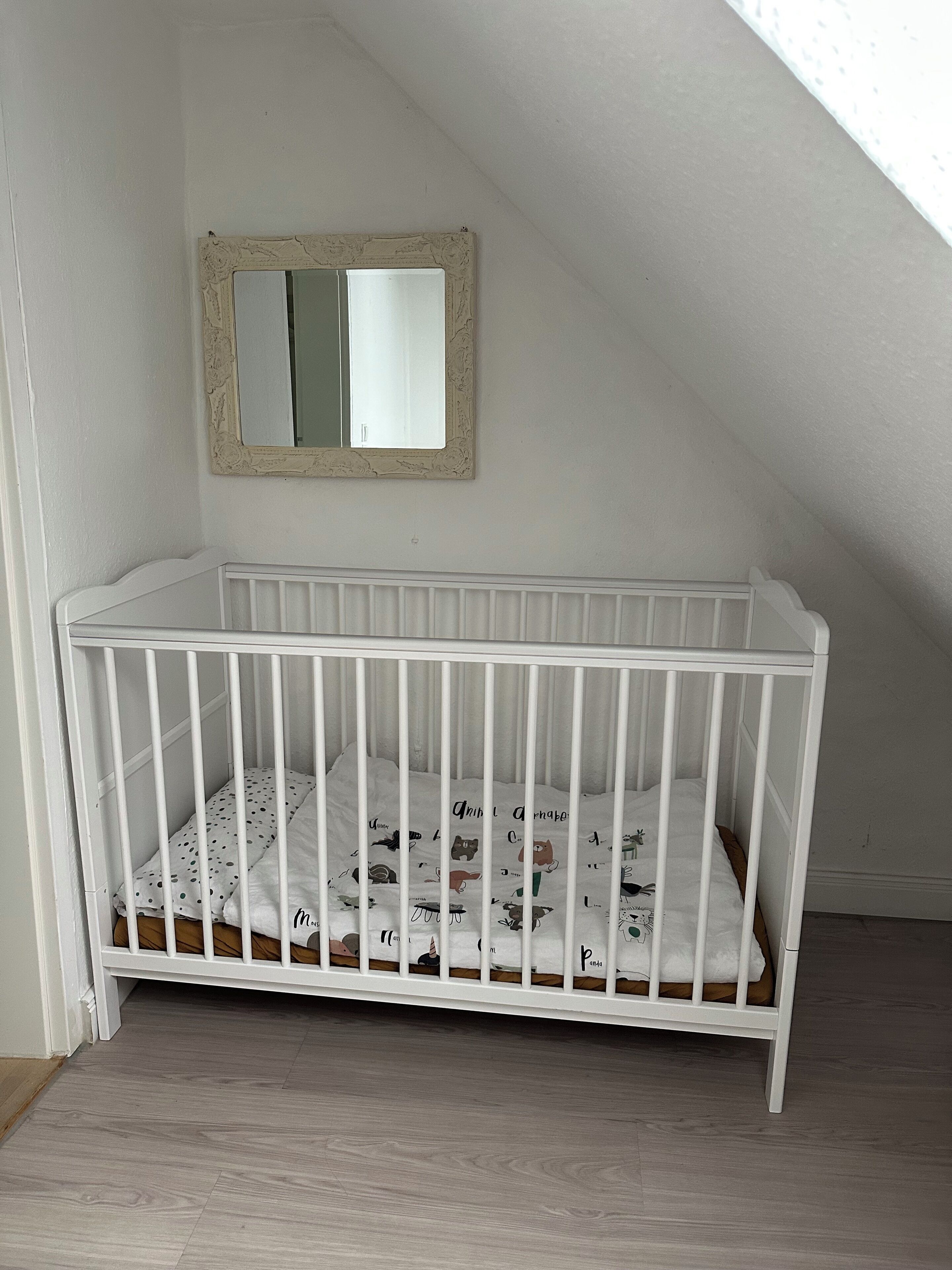 Een wit kinderbed met een stippenmatras in een kamer met schuine wanden en een kleine spiegel aan de muur.