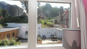 Blick aus einem Fenster auf einen Raum mit einer Holzterrasse, Gartenmöbeln und einem großen Topfpflanzen.