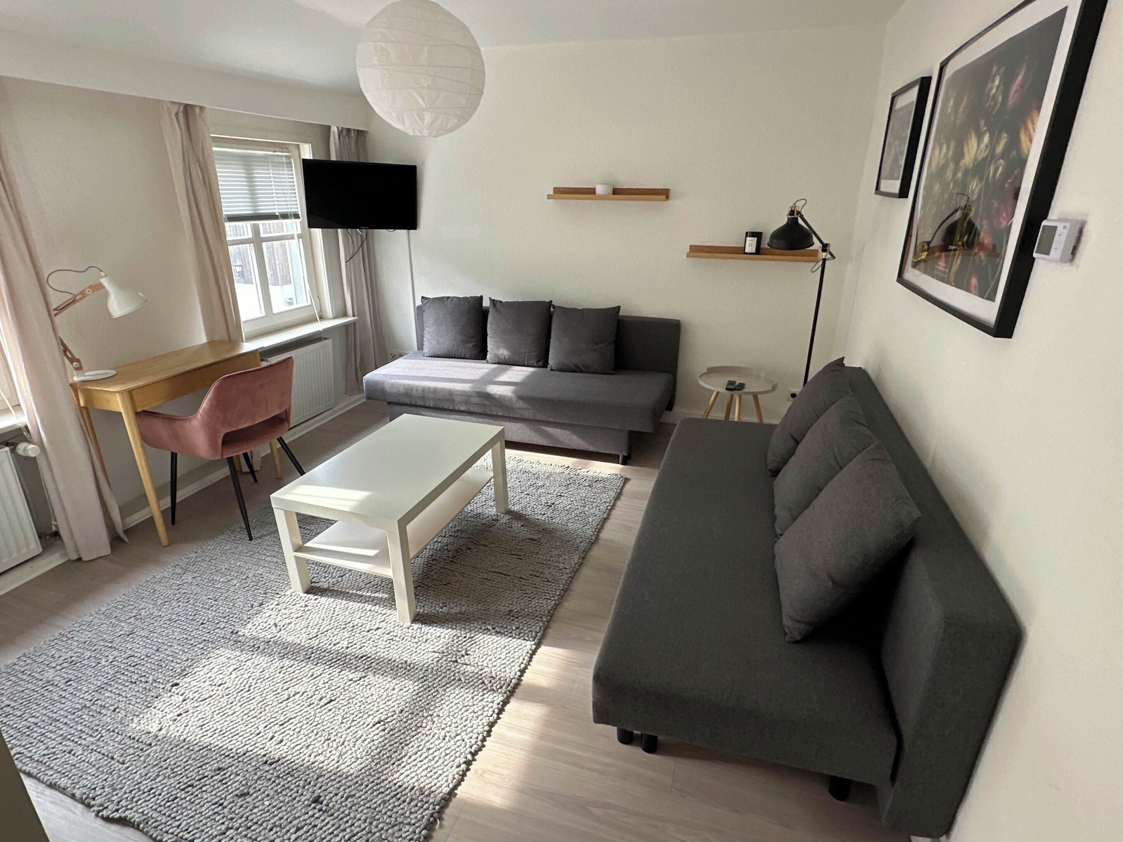Ett modernt vardagsrum med en grå soffa, ett vitt soffbord, ett skrivbord i trä och en tv som är monterad på väggen.