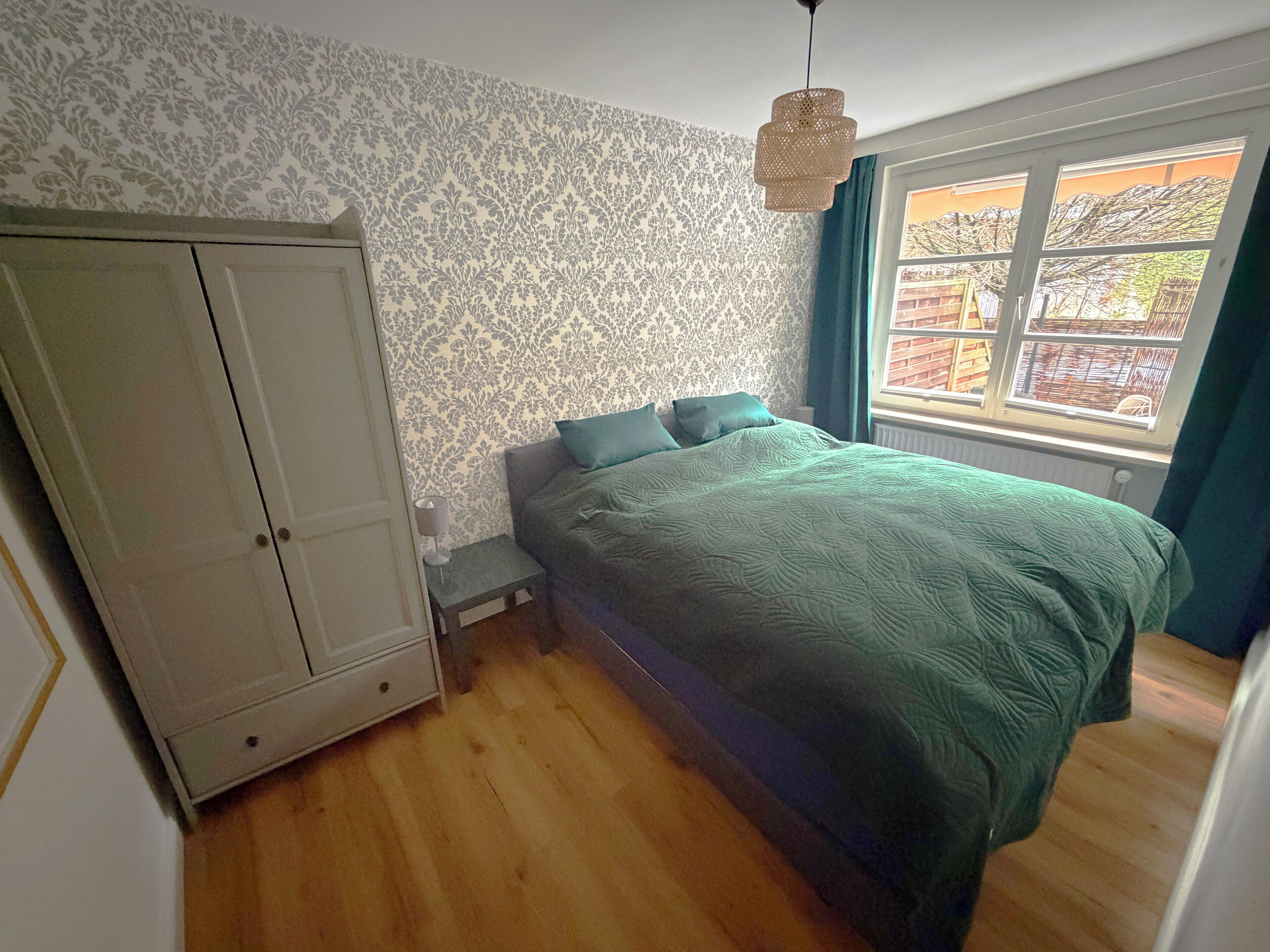A bedroom with a bed, a wardrobe, a nightstand, a lamp, and a window with curtains.