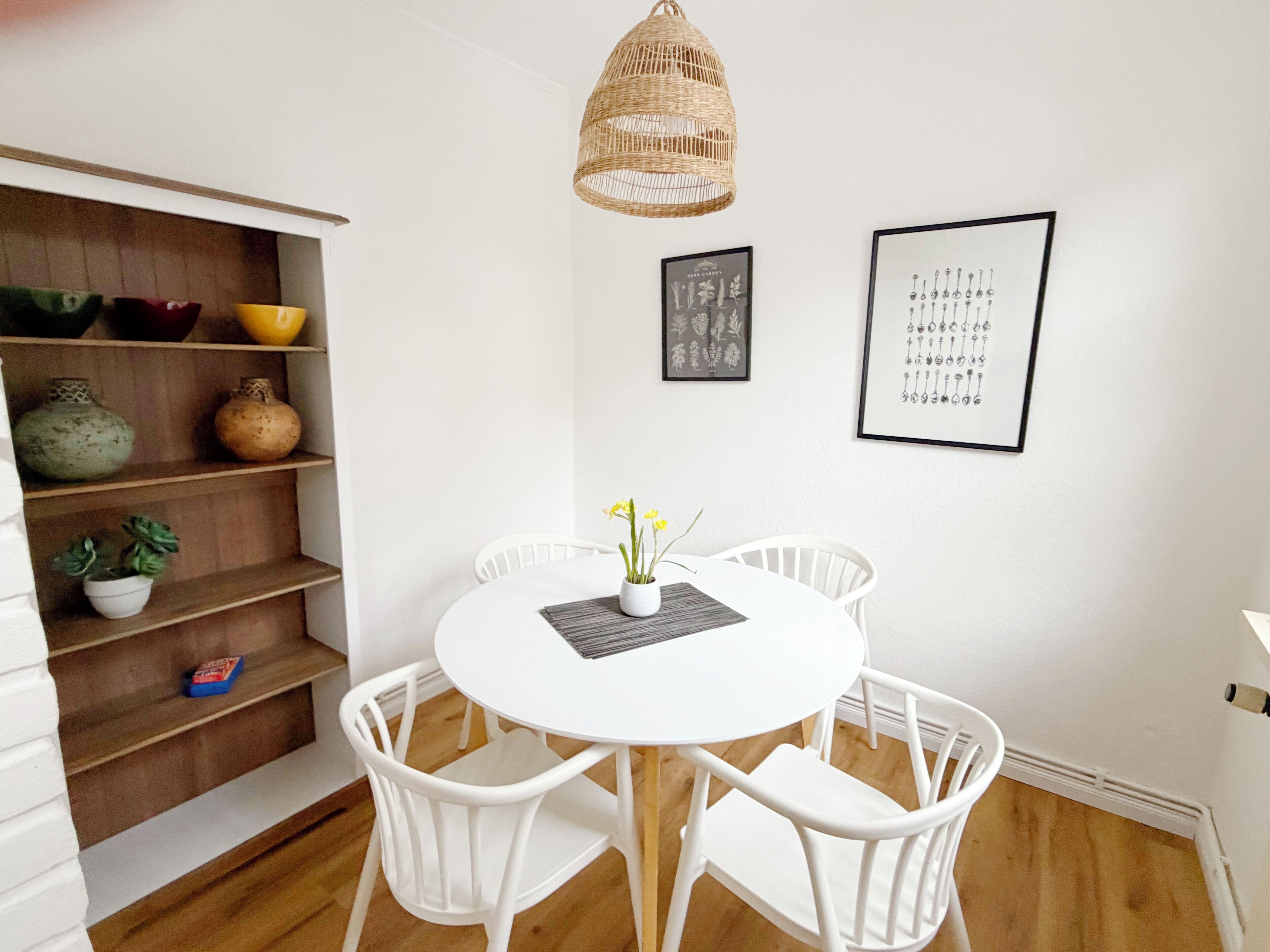 A dining area with a round white table, four white chairs, a woven pendant light, and a shelf with decorative items.