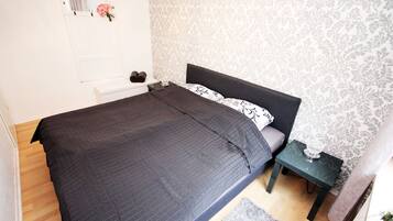 Een slaapkamer met een donker bed, een klein nachtkastje met een lamp, en behang met een patroon.