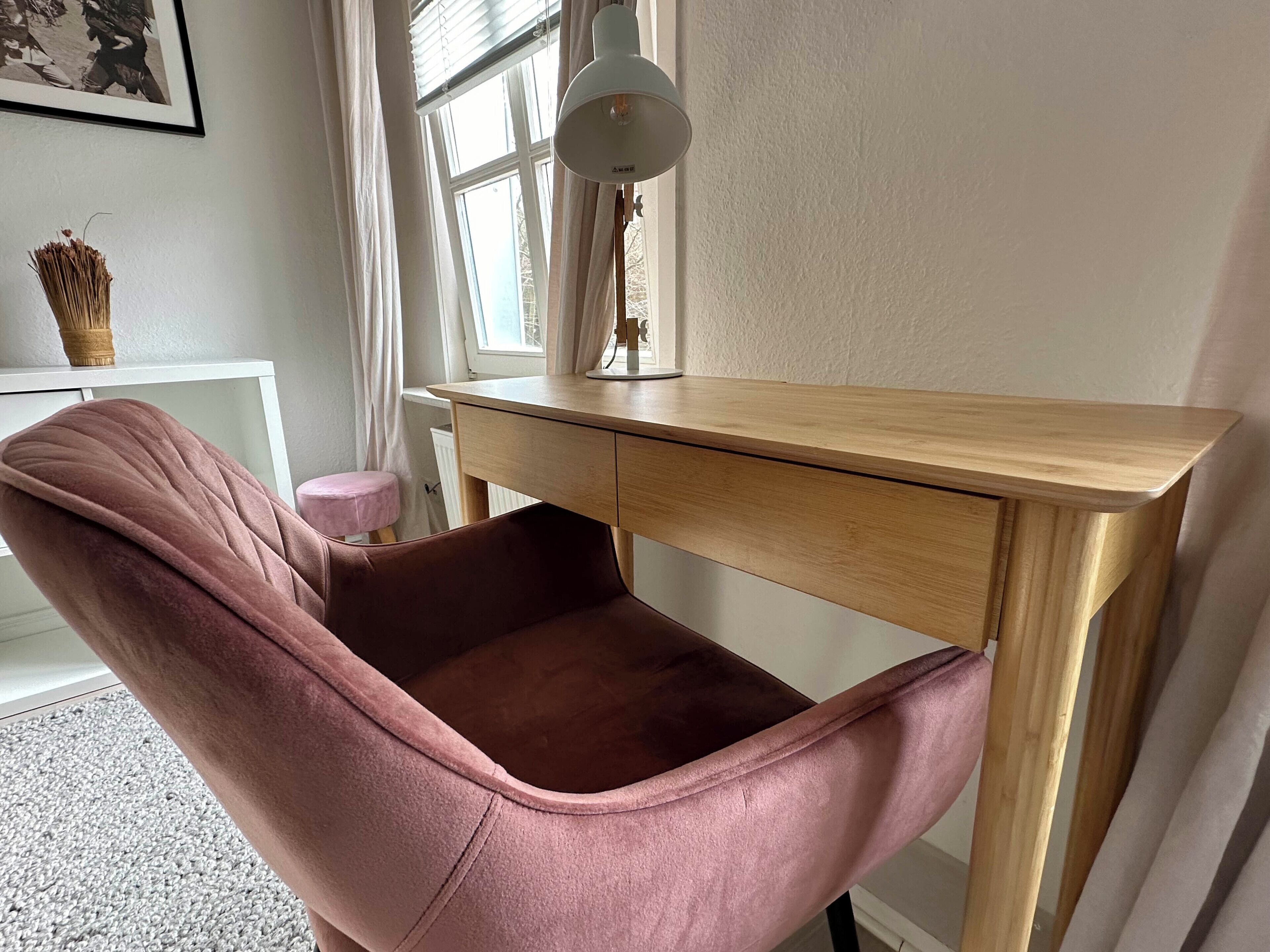 Une pièce avec un bureau en bois, une chaise rose, un radiateur blanc et une fenêtre avec des rideaux.