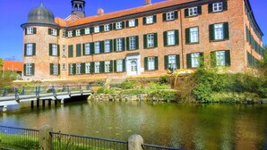 Ein großes Backsteingebäude mit einer zentralen Kuppel und einem Glockenturm, an einem ruhigen Fluss gelegen, über den eine Brücke führt.