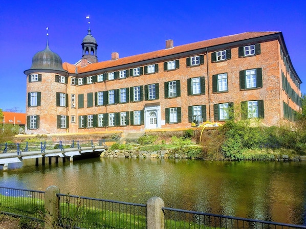 Un gran edificio de ladrillo con una cúpula central y un campanario, ubicado junto a un río tranquilo con un puente.