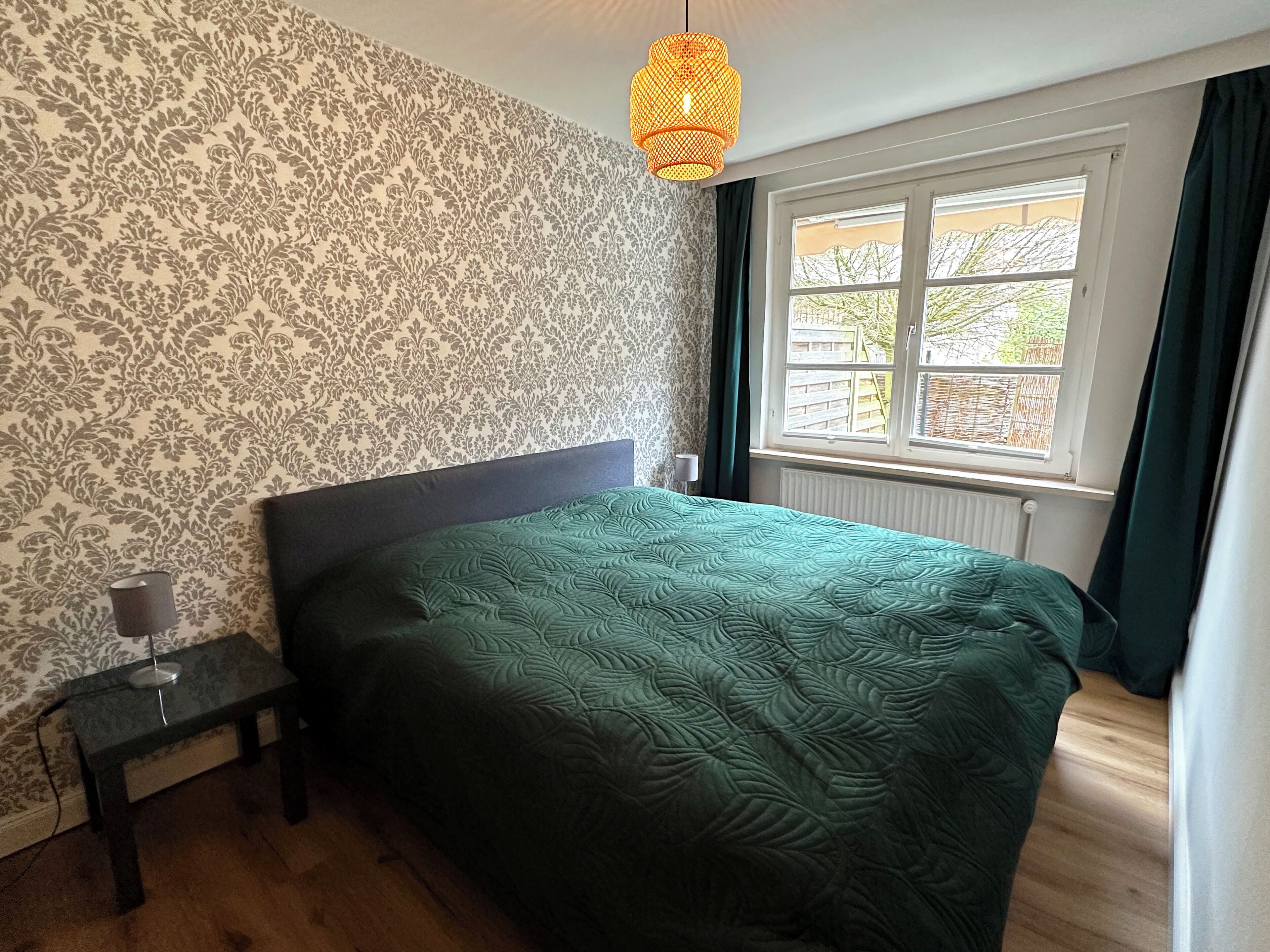 Ein Schlafzimmer mit strukturiertem Wandpapier, einer grünen Bettdecke, einem Nachttisch mit Lampe und einem Fenster mit Vorhängen.
