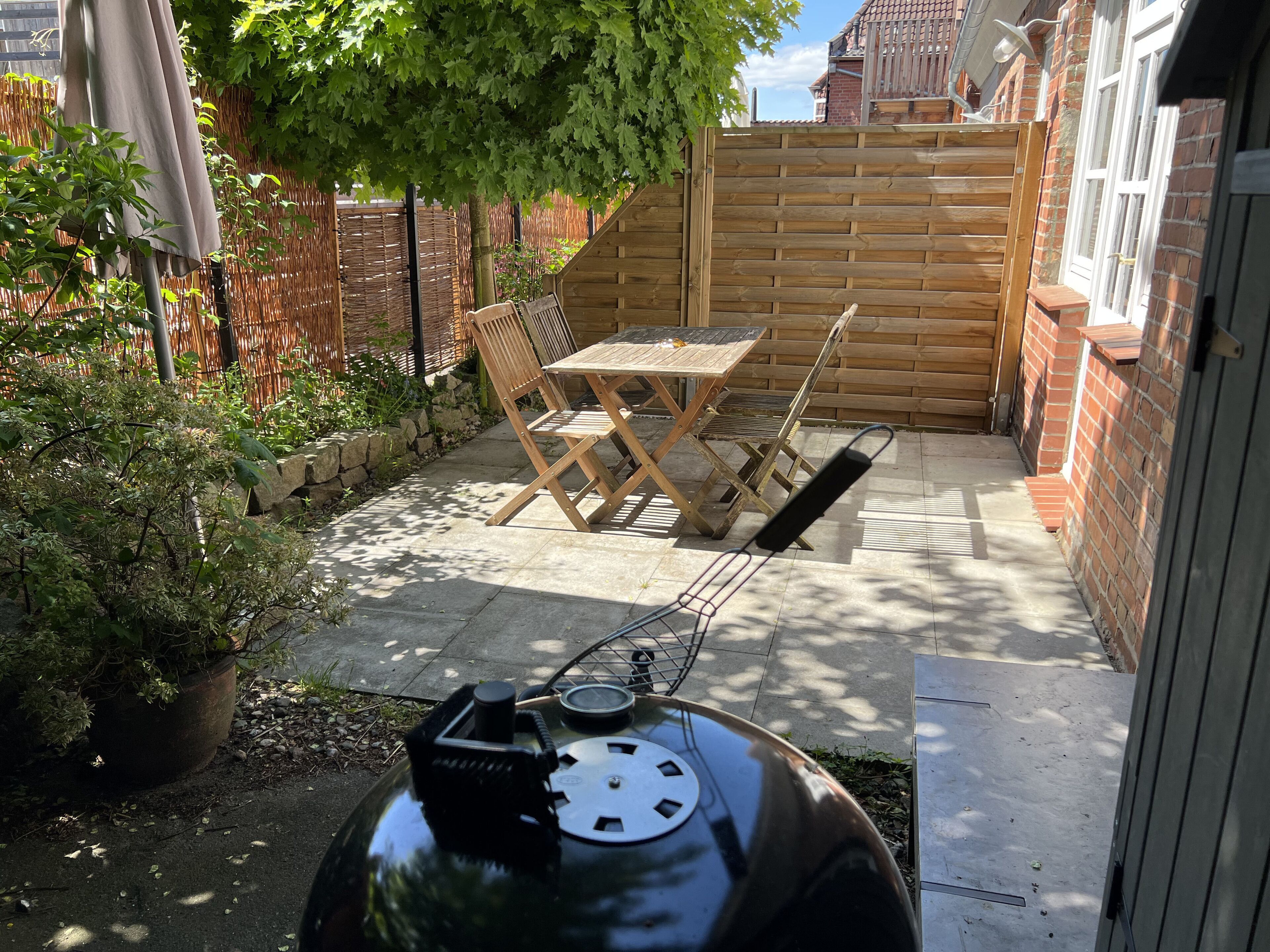 Un patio con una parrilla, mesa y sillas de madera, una planta en maceta y una cerca de bambú.